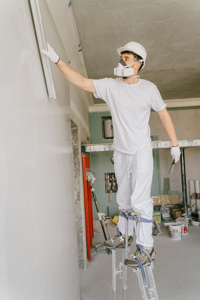 Crafting Captivating Headlines: Your awesome post title goes here Construction worker on stilts painting a wall indoors, wearing protective gear and a hard hat.