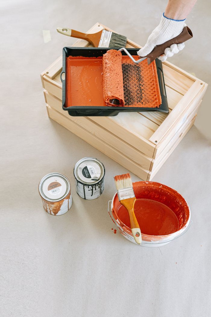Flat lay of painting supplies on wooden crate, including roller, brushes, and paint cans.